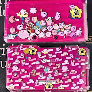 May include: A pink Nintendo 3DS case decorated with numerous Kirby characters in different poses. The case includes the text "Kirby 30th Anniversary" in a yellow emblem, marking the franchise's 30th anniversary.