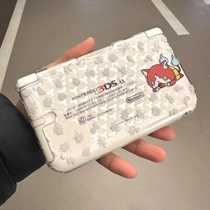 Puede incluir: Consola de juegos portátil Nintendo 3DS LL blanca con una funda protectora transparente. La funda presenta un patrón de siluetas de personajes grises y un personaje de dibujos animados rojo y amarillo. La marca de la consola es visible.