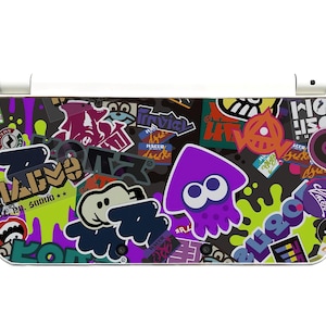May include: A Nintendo 3DS console skin with a vibrant design of cartoon graphics and text. The skin features a purple squid, green paint splatters, and sticker-like images in multiple colours, creating a playful aesthetic.