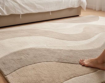 May include: A neutral-toned area rug with a wavy pattern in shades of beige, tan, and gray. The rug is placed on a hardwood floor in a bedroom setting, with a bed and a foot visible.