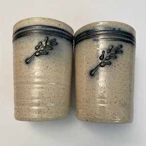 May include: Two ceramic mugs with a speckled glaze and a blue and silver design. Each mug features a stylized sprig of leaves.