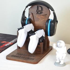 Könnte beinhalten: Ein dunkler Holzständer für Gaming-Headsets und Controller. Er zeigt ein schwarz-blaues Headset und zwei weiße Controller. Der Ständer trägt den Text "DAD BY DAY GAMER BY NIGHT". Eine kleine Astronautenfigur und ein schwarzer Controller sind ebenfalls vorhanden.
