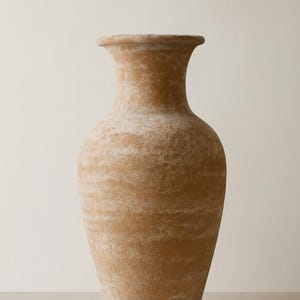 May include: A tall, terracotta-colored vase with a wide, rounded body and a flared rim. The surface has a textured, aged appearance with lighter, mottled areas, suggesting a weathered finish. The vase is suitable for home decor.