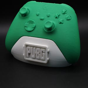 Könnte beinhalten: Ein grüner und weißer Controller mit dem Wort "PUBG" auf der Unterseite. Der Controller hat Tasten und Joysticks und ist für Spiele konzipiert. Der Controller ist aus Kunststoff.