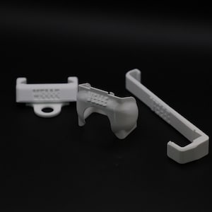 Op de afbeelding: Drie witte 3D-geprinte componenten worden getoond tegen een donkere achtergrond. De stukken omvatten een beugel met tekst, een gebogen stuk en een lang, rechthoekig stuk met een haak.