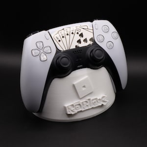 Pode incluir: Controlo PlayStation 5 branco e preto com um design de cartas de jogar, repousando sobre um suporte branco com o logótipo Roblox. O controlo apresenta um centro preto e botões brancos. O design das cartas de jogar inclui espadas.