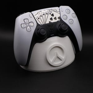 May include: A white and black PlayStation 5 controller with a playing card design, resting on a matching white stand. The controller features black joysticks and buttons, with a spades-themed card design on the touchpad. The stand has a circular design.