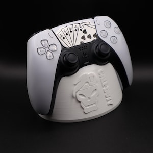 Pode incluir: Um controle PlayStation 5 branco e preto com um design de cartas de baralho no touchpad, apoiado em um suporte cinza. O suporte apresenta um gráfico de caveira e a palavra "Call of Duty".