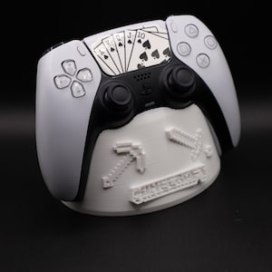 May include: A white and black PlayStation 5 controller with a playing card design on the touchpad, resting on a white stand. The stand features pixelated graphics and the word "Minecraft".