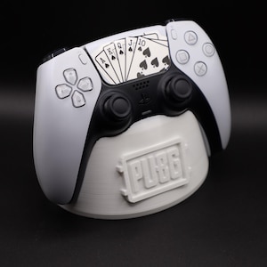 May include: White and black PlayStation 5 controller with a playing card design, resting on a white stand with the PUBG logo. The controller features a black touchpad and buttons, with a fan of spades cards on the right side.