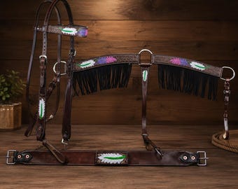 May include: A dark brown leather horse bridle and breast collar set featuring black fringe and hand-painted feather motifs in white, green, and purple. The tack includes silver-toned metal buckles, rings, and hardware throughout the assembly.