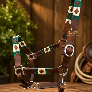 May include: A brown leather horse halter featuring woven fabric accents in cream, dark green, and brown geometric patterns. The halter includes silver-toned metal hardware, buckles, and rings, with the text THE SADDLE TRUNK printed on the side strap.