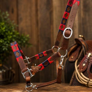 May include: A brown leather horse halter featuring woven fabric accents in red and navy blue with a geometric diamond pattern. The halter includes silver-toned metal buckles, rings, and a snap hook, with the text THE SADDLE TRUNK printed in white.