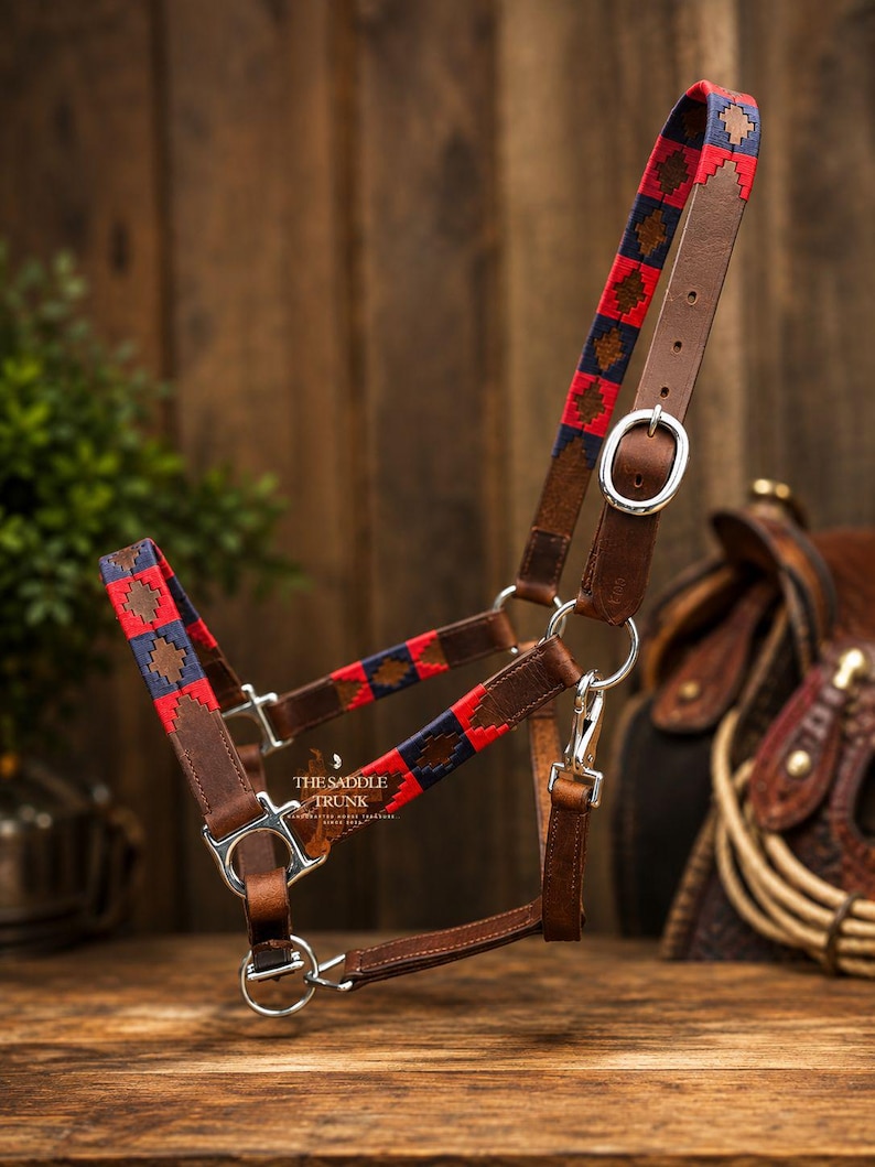 May include: A brown leather horse halter featuring woven fabric accents in red and navy blue with a geometric diamond pattern. The halter includes silver-toned metal buckles, rings, and a snap hook, with the text THE SADDLE TRUNK printed in white.