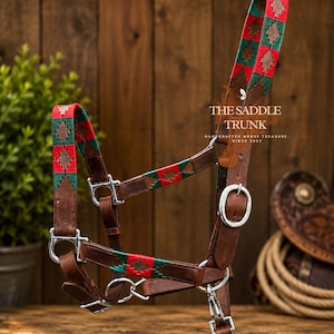 May include: A brown leather horse halter featuring woven fabric accents in red and green with a diamond pattern. The halter includes silver-colored metal hardware, buckles, and a snap hook. Text on the leather reads THE SADDLE TRUNK HANDCRAFTED HORSE TREASURE SINCE 2022.