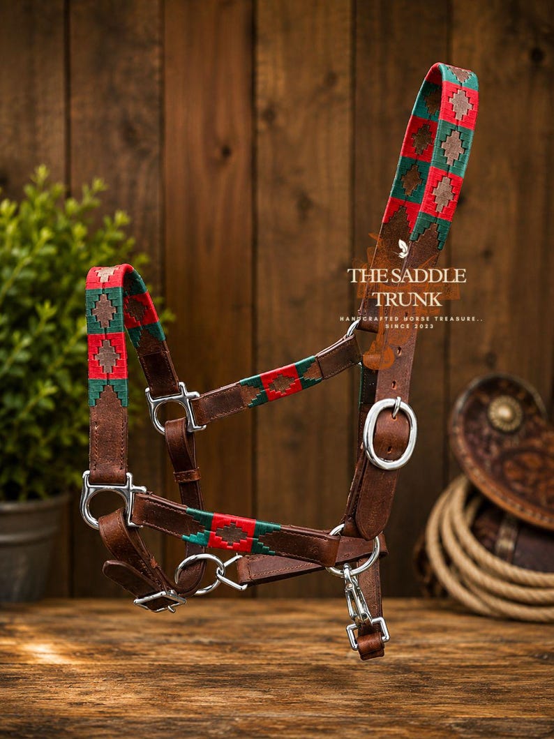 May include: A brown leather horse halter featuring woven fabric accents in red and green with a diamond pattern. The halter includes silver-colored metal hardware, buckles, and a snap hook. Text on the leather reads THE SADDLE TRUNK HANDCRAFTED HORSE TREASURE SINCE 2022.