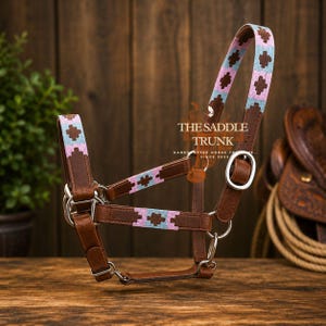 May include: A brown leather horse halter featuring woven fabric accents in light blue and pink with a geometric cross pattern. The halter includes silver-toned metal hardware, buckles, and rings. The text THE SADDLE TRUNK is visible on the side.