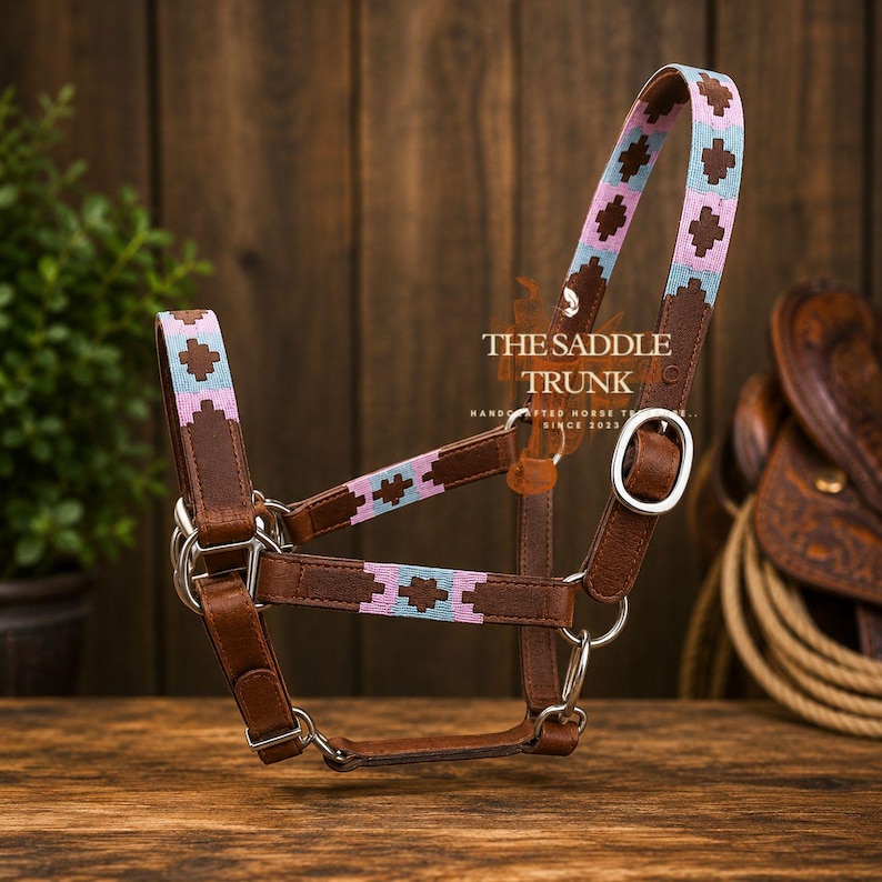 May include: A brown leather horse halter featuring woven fabric accents in light blue and pink with a geometric cross pattern. The halter includes silver-toned metal hardware, buckles, and rings. The text THE SADDLE TRUNK is visible on the side.