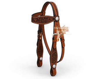 May include: A tan leather horse headstall featuring intricate floral tooling and dark brown accents. The headstall includes silver-toned metal conchos and buckles, with the text THE SADDLE TRUNK visible in the center. The design showcases detailed craftsmanship on the browband and cheek pieces.