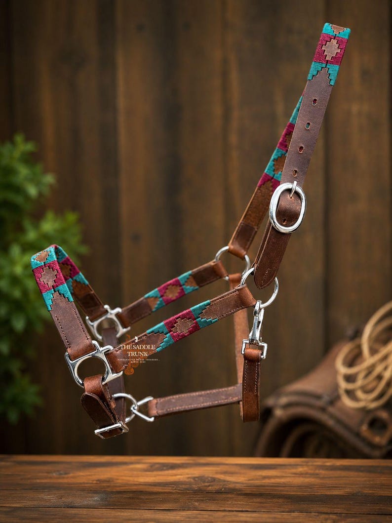 May include: A brown leather horse halter featuring woven fabric accents in teal and magenta with a geometric diamond pattern. The halter includes silver-toned metal buckles, rings, and clips, with the text THE SADDLE TRUNK printed on the leather.