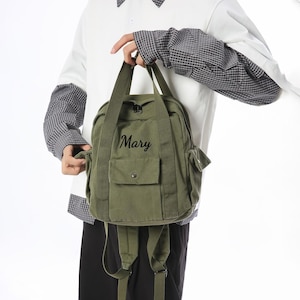 May include: An olive green backpack with a top handle and shoulder straps. The backpack features a front pocket with a button closure and the name "Mary" in script. The bag is made of a canvas-like material.