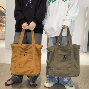 May include: Two canvas tote bags, one mustard yellow and one olive green, are held by people. The mustard bag has a black "S" embroidered on the front pocket. The olive green bag has a black "K" embroidered on the front pocket. Both bags have long handles.