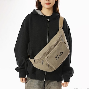 May include: A khaki-colored fanny pack with a front pocket and a zipper closure. The name "Linda" is written in cursive on the front of the bag. The bag has a tan strap and is worn across the body.