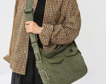 May include: Olive green messenger bag with two front pockets and a flap closure. The bag has a shoulder strap and a distressed patch detail. The letter "M" is embroidered on the flap. The bag is from XZminimalistBag.