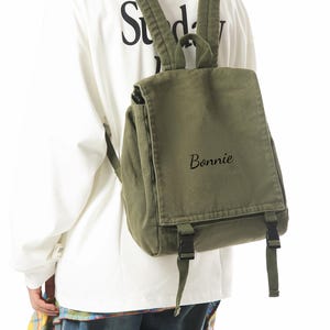 May include: An olive green backpack with adjustable straps and a flap closure. The word "Bonnie" is written on the front. The person in the image is wearing a white long-sleeve shirt with the word "Sunday" printed on the back.