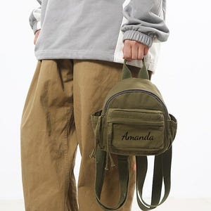 May include: An olive green mini backpack with the name "Amanda" embroidered on the front. The backpack has a small top handle, two side pockets, and adjustable straps. The person is wearing a grey sweatshirt and tan trousers.