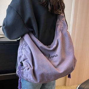 Puede incluir: Un bolso de hombro de color lavanda con el nombre "Anna" bordado en la parte delantera. El bolso tiene un cierre de cordón, un pequeño bolsillo lateral y una borla morada. El bolso se lleva sobre una camiseta negra y unos vaqueros azules.