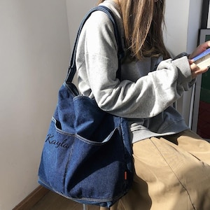 May include: A dark blue denim tote bag with two front pockets and a shoulder strap. The name "Kayla" is embroidered on the front. The bag has a rectangular shape and a small orange tag on the side. The text "XZminimalistBag" is at the bottom.