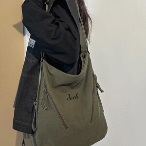 May include: Olive green canvas shoulder bag with two zippered pockets and a long adjustable strap. The word "Jack" is printed on the front. The bag is part of the XZminimalistBag collection.