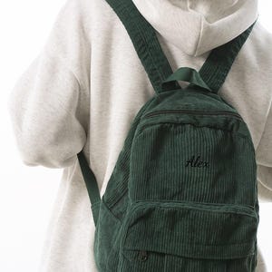 Puede incluir: Una mochila de pana verde oscuro con el nombre "Alex" bordado en la parte delantera. La mochila tiene un bolsillo frontal, un asa superior y correas ajustables para los hombros. La bolsa se lleva sobre una sudadera con capucha gris claro.