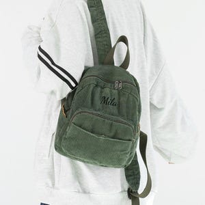 Könnte beinhalten: Ein grüner Cord-Rucksack mit einer Vordertasche und einem oberen Griff. Auf dem Rucksack ist der Name "Mila" aufgestickt. Die Tasche wird über der Schulter getragen.