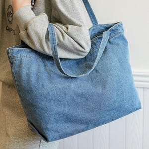 May include: A large, light blue denim tote bag with two shoulder straps. The bag is rectangular with a wide opening and a simple design. The bag is held by a person wearing a gray sweatshirt. The text "X ZminimalistBag" is at the bottom right.