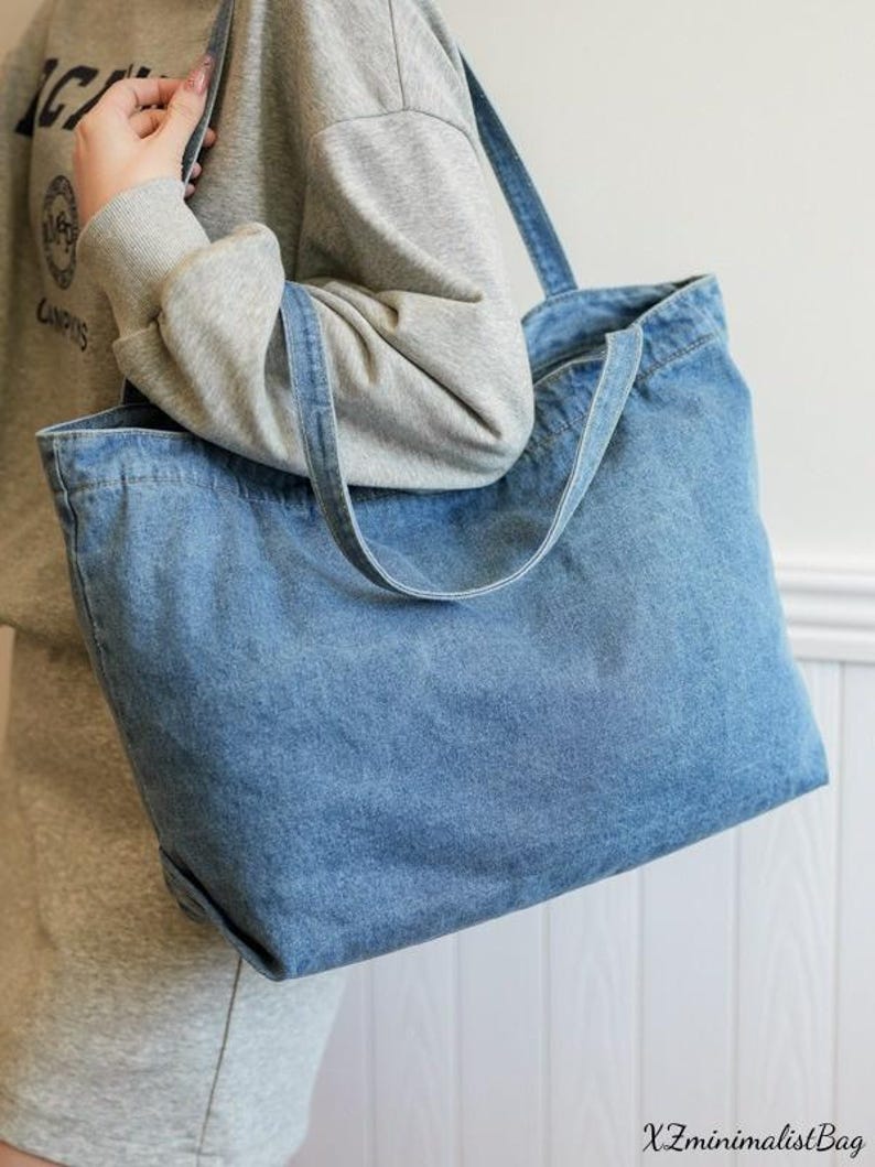 May include: A large, light blue denim tote bag with two shoulder straps. The bag is rectangular with a wide opening and a simple design. The bag is held by a person wearing a gray sweatshirt. The text "X ZminimalistBag" is at the bottom right.