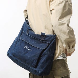 May include: A dark blue denim tote bag with a front pocket and the name "Ethan" embroidered in white. The bag has a shoulder strap and is worn over the shoulder. The bag is part of a minimalist bag collection.