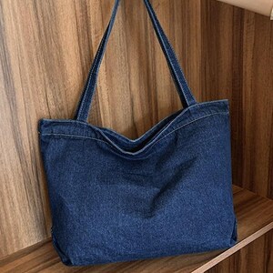 May include: A dark blue denim tote bag with long straps and white stitching. The bag is held up by a hand, showcasing its shape and design. The bag is a simple, yet stylish accessory.