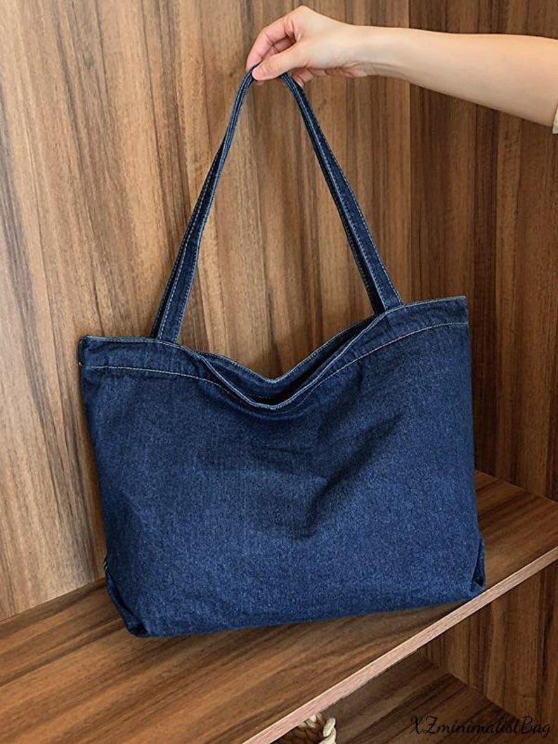 May include: A dark blue denim tote bag with long straps and white stitching. The bag is held up by a hand, showcasing its shape and design. The bag is a simple, yet stylish accessory.