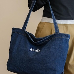 May include: A dark blue denim tote bag with long straps. The word "Amelia" is embroidered in white script on the front of the bag. The bag is rectangular and appears to be made of a sturdy denim material.