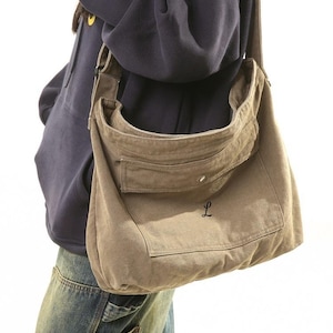 Op de afbeelding: Een olijfgroene canvas messenger bag met een grote voorzak en een knoopsluiting. De letter "L" is op de voorkant geborduurd. De tas wordt over de schouder gedragen. De tekst "XZminimalistBag" staat onderaan.