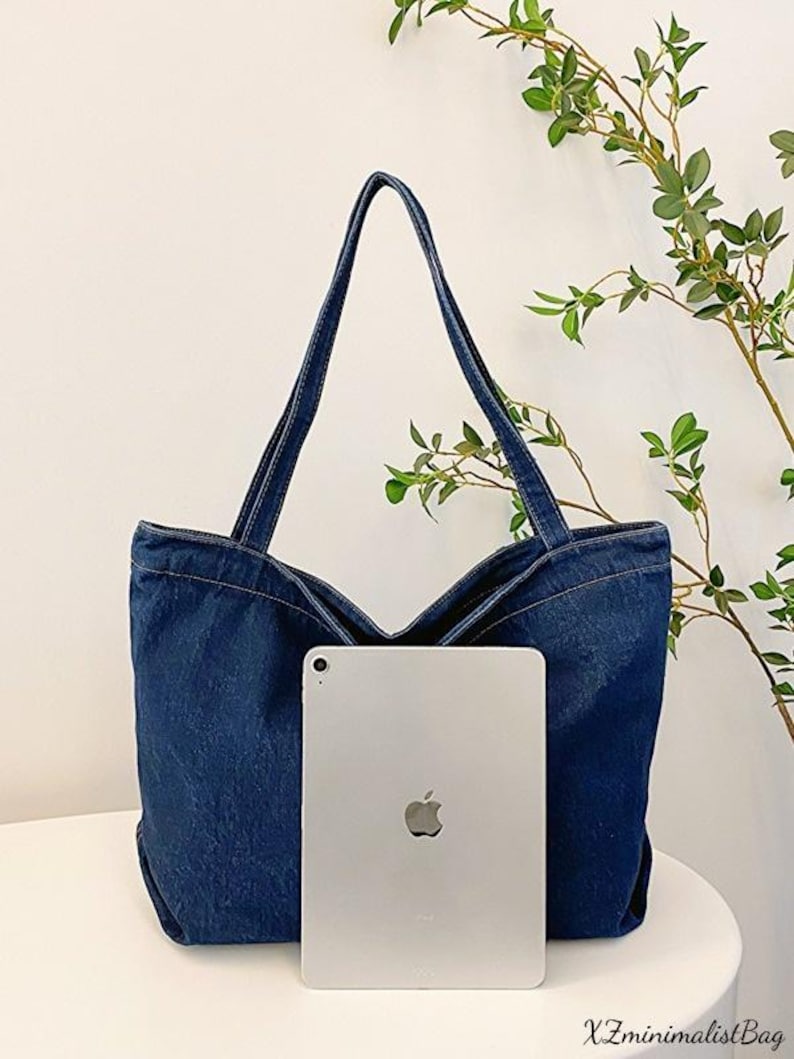 May include: A dark blue denim tote bag with long straps. The bag has a wide opening and is shown with a silver tablet in front. The bag is sitting on a white surface. The text "XZminimalistBag" is at the bottom.