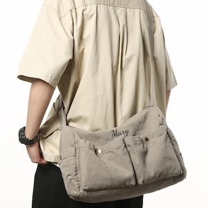 Puede incluir: Bolso mensajero de lona beige con múltiples bolsillos delanteros y una correa para el hombro. La palabra "Mary" está bordada en el bolso. El bolso se lleva al hombro.