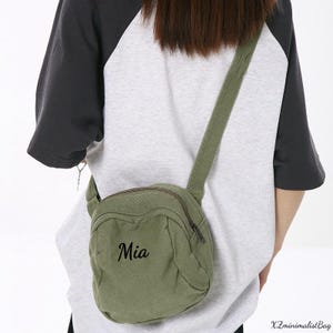 May include: Olive green crossbody bag with a zippered main compartment and adjustable strap. The bag features the name "Mia" in black script. The bag is worn over a gray and black raglan sleeve shirt.