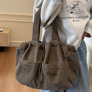 Op de afbeelding: Een bruine corduroy tote bag met twee voorzakken en een trekkoordsluiting. De tas heeft twee handvatten en de naam "Helen" geborduurd op een van de zakken. De tas heeft een neutrale kleur en lijkt een casual stijl te hebben.