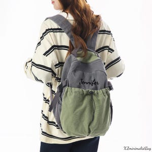 May include: A gray and green canvas backpack with the name "Jennifer" printed on the front. The backpack has adjustable straps and a drawstring closure on the front pocket. The bag is worn by a person wearing a striped shirt.