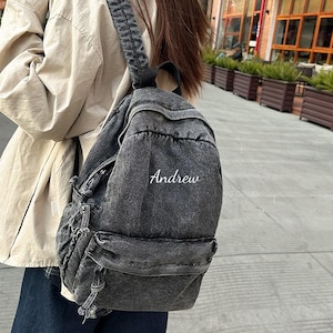 May include: A gray denim backpack with the name "Andrew" embroidered in white. The backpack has adjustable straps and multiple pockets. The bag is worn over the shoulder.