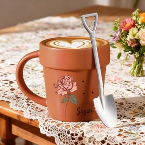 Può includere: Una tazza color terracotta a forma di vaso, con un'illustrazione di rosa rosa e il nome "Susan". La tazza contiene un latte con un disegno a forma di cuore, accompagnato da un cucchiaio argentato a forma di pala. Un mazzo di fiori è nelle vicinanze.