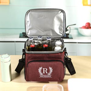 May include: Maroon insulated cooler bag with a black shoulder strap, open to reveal bottles and small containers. The bag has a front pocket with the text "R RICHARD" in a white decorative font. The interior is silver.
